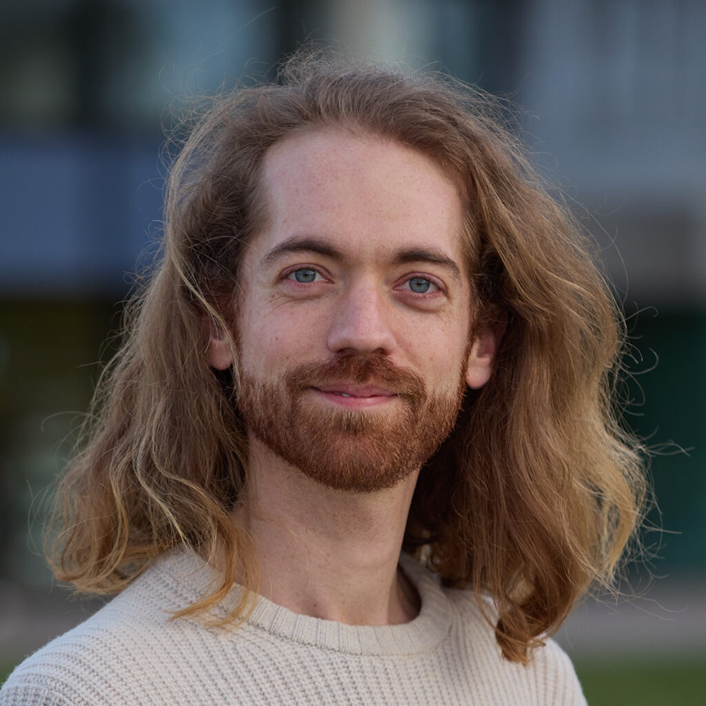 ein Porträt eines weissen Manns mit langen, hellen Haaren und Bart. Im Hintergrund verschwommen Häuser und grün