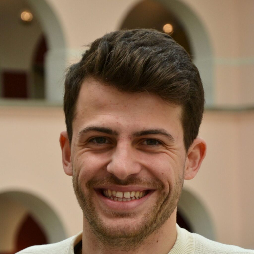 Portrait eines weissen Mannes mit kurzen, dunken Haaren und 3-Tage-Bart. Er lächelt in die Kamera, im Hintergrund ein Gebäude mit Bogenfenstern