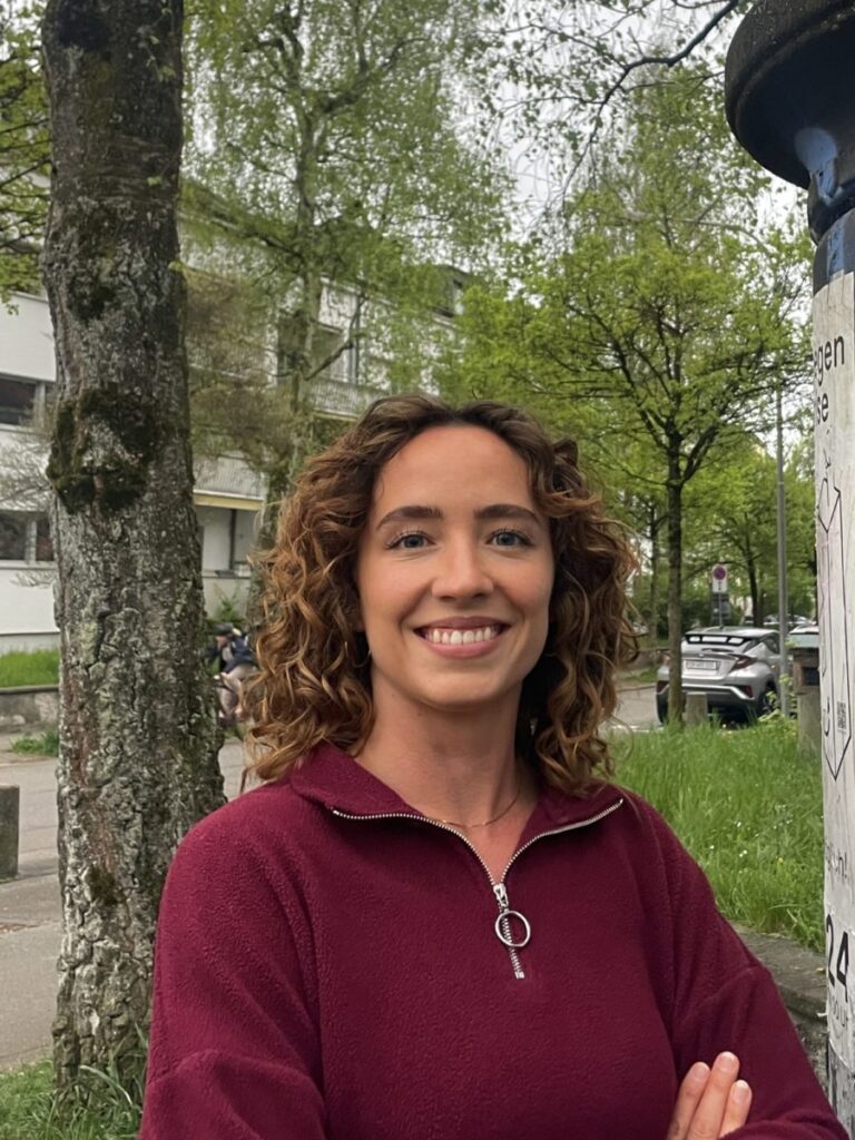 Frontalportrait einer weissen Frau mit gelockten, halblangen Haaren. Sie lächelt strahlend in die Kamera und steht vor einer sehr begrünten Quartierstrasse