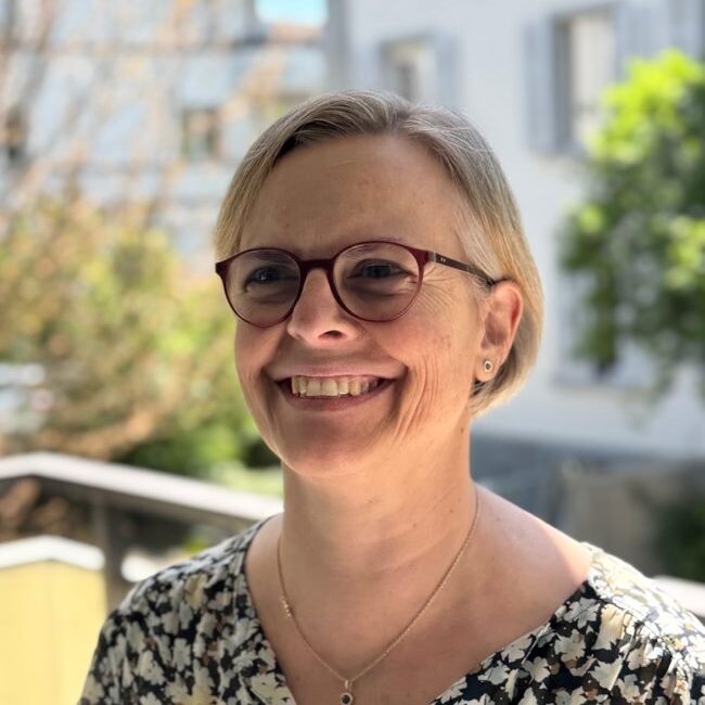 Portrait einer weissen Frau mit kurzen, blonden Haaren und einer Brille. Sie lächelt jemanden neben der Kamera an. Hinter ihr Häuser und Grün einer Stadt